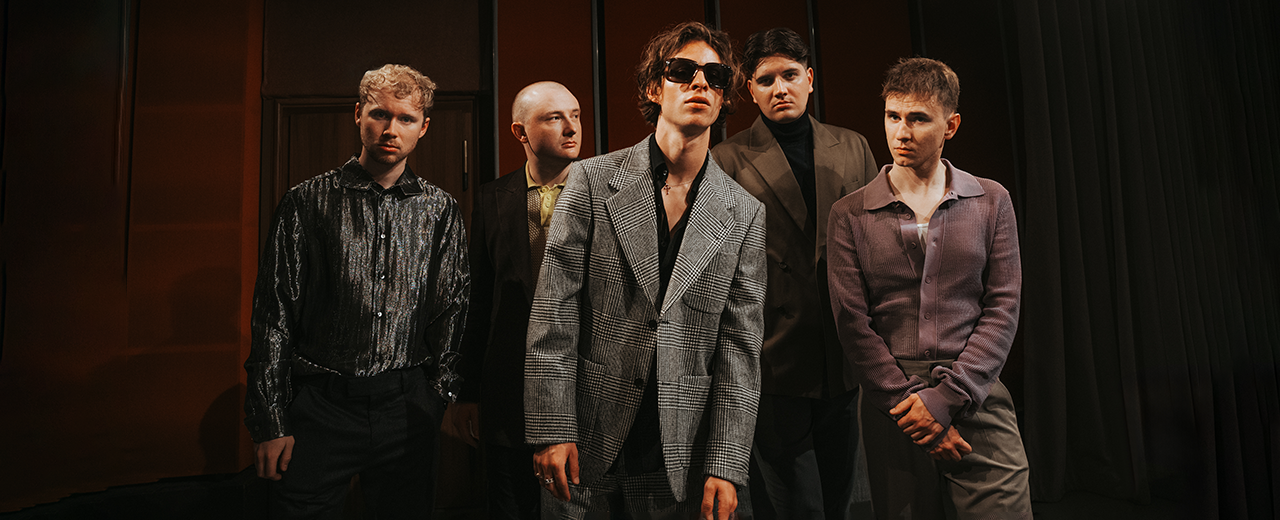 Fünfköpfige Indie-Band, Frontmann im grauen karierten Anzug mit Sonnenbrille, die Gruppe posiert in einem abgedunkelten Raum.