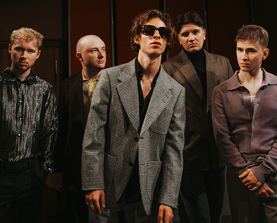 Fünfköpfige Indie-Band, Frontmann im grauen karierten Anzug mit Sonnenbrille, die Gruppe posiert in einem abgedunkelten Raum.